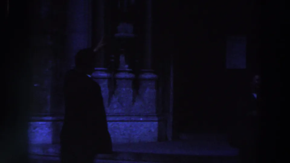 A dark-suited man in 1961 Ireland footage gestures upward beside a blue-lit stone building exterior.