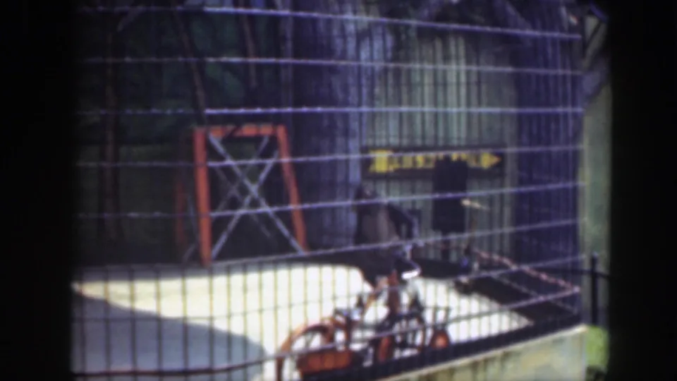 The child on the bicycle passes low in frame inside the fenced 1948 Washington, DC play area, partly obscured by mesh and shadow.