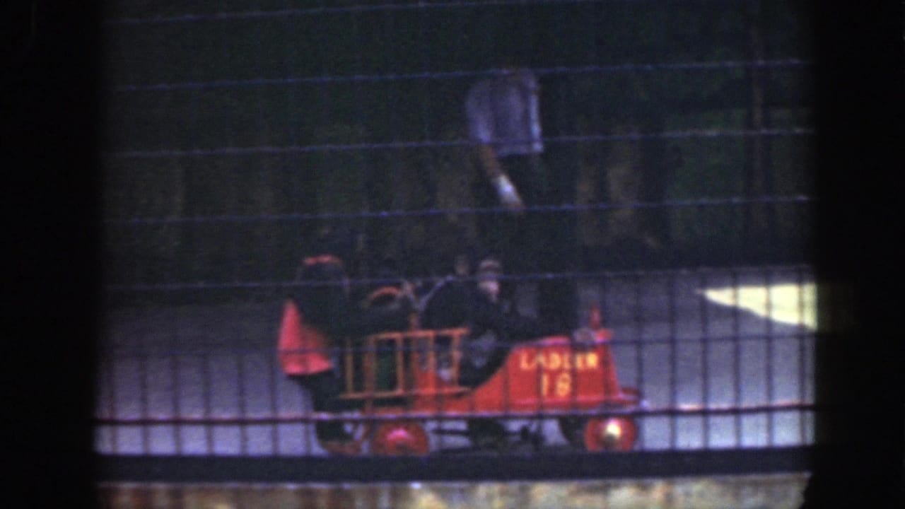 Two chimpanzees ride a red toy fire truck behind a wire-mesh zoo fence.