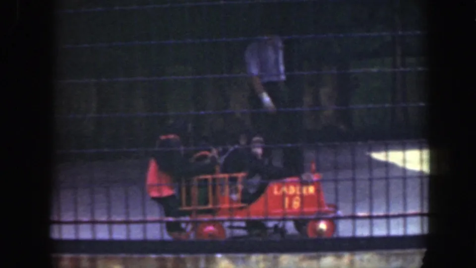 1948 Washington DC zoo footage shows two chimpanzees in a red toy fire truck behind wire mesh, with enclosure fencing in the background.