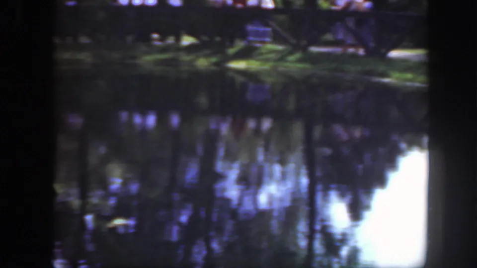 1948 Washington DC home-movie frame showing a pond, tree reflections, a wooden fence, and distant people along a grassy bank.