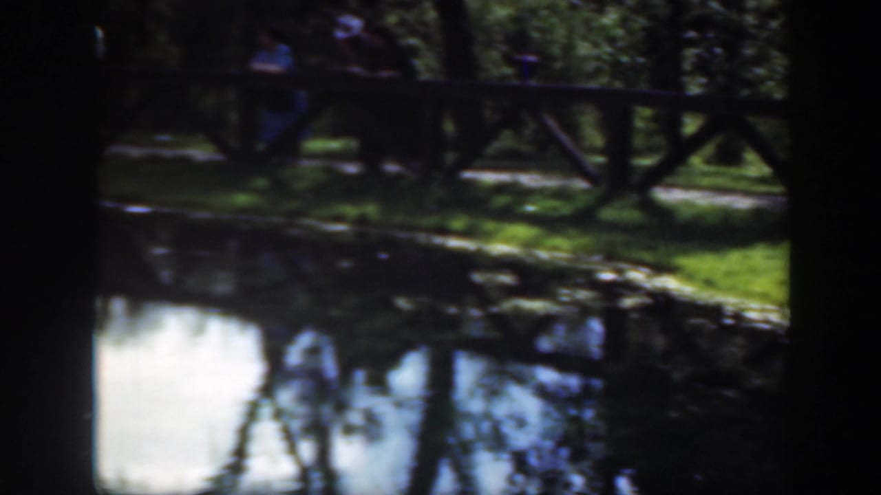 1948 Washington DC home-movie frame of a shaded pond edge, wooden fence, grass, and distant park visitors near a path.