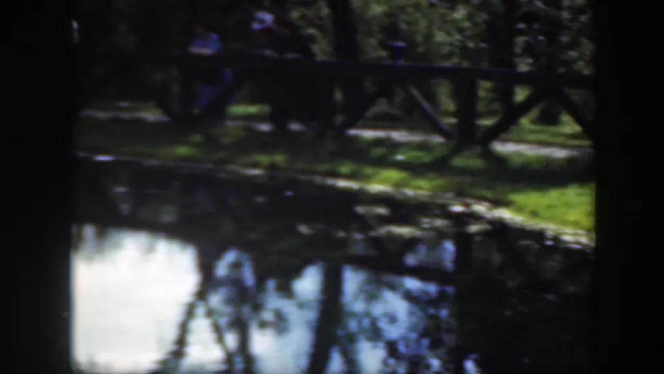 1948 Washington DC home-movie frame of a shaded pond edge, wooden fence, grass, and distant park visitors near a path.