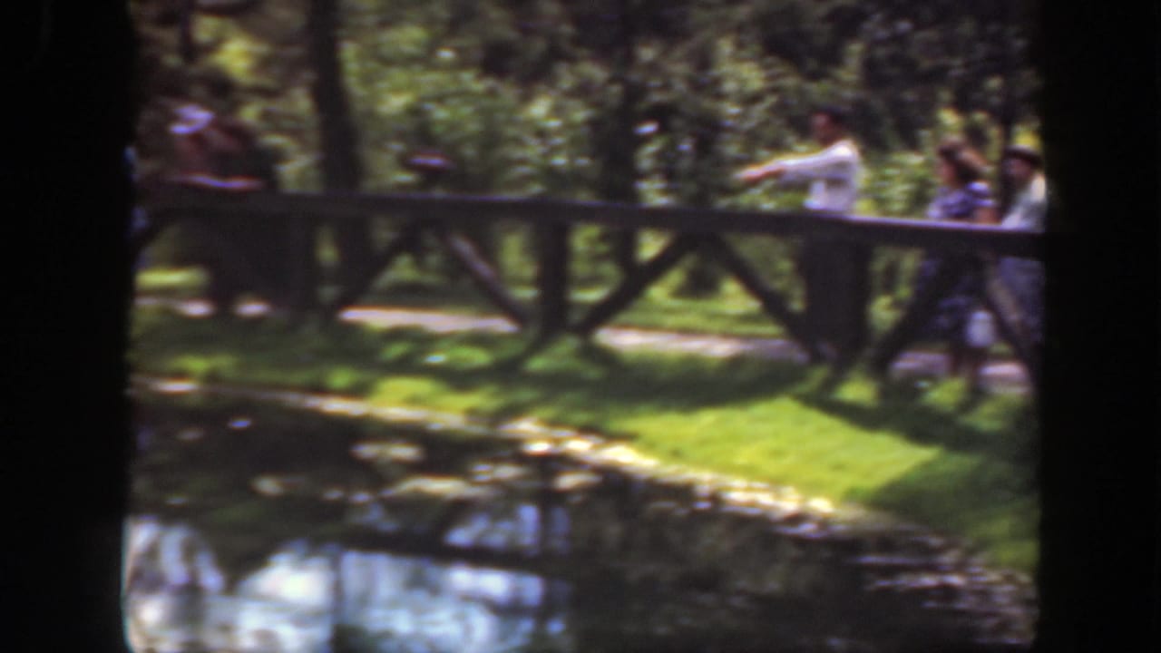 1948 Washington DC park frame showing people near a wooden fence, a pond edge, grass, trees, and bright reflected water.