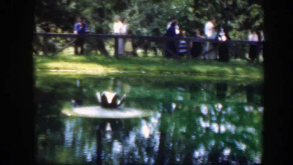 1948 Washington, DC frame with adults in period clothing on a wooden bridge over or beside a landscaped pond.