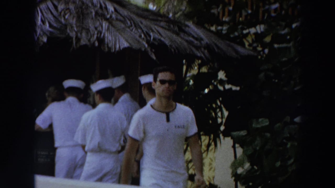 Men in white sailor-style uniforms gather beside a thatched hut while a man in a white shirt and sunglasses passes in the foreground.