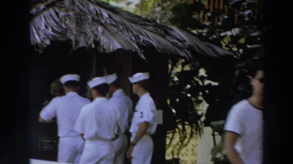1966 Puerto Rico resort scene with uniformed men beneath a thatched roof and a man in a white shirt moving out of view.