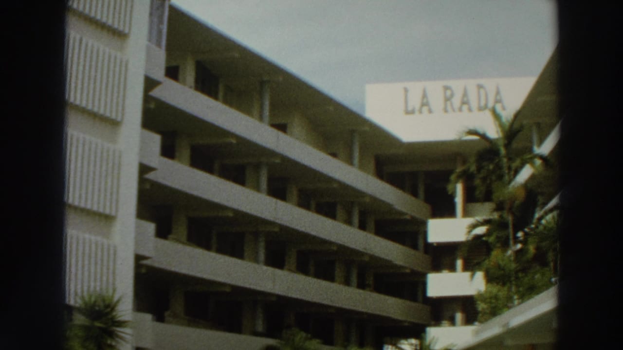 White multi-story building with balconies and a La Rada sign behind palm foliage.