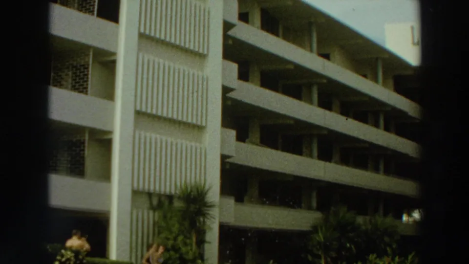1966 Puerto Rico frame showing the building corner, stacked balconies, vertical panels, tropical landscaping, and a small visible person near the bottom.