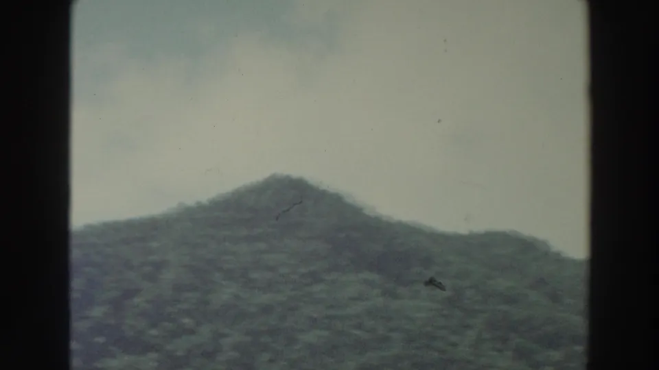 1966 Puerto Rico home-movie frame of a rounded tree-covered hillside below a muted, lightly clouded sky.