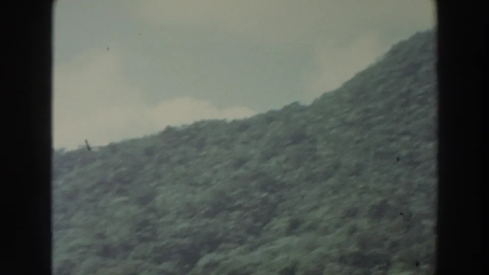 1966 Puerto Rico 8mm frame showing a close sloping wooded hillside under a pale sky with light cloud cover.