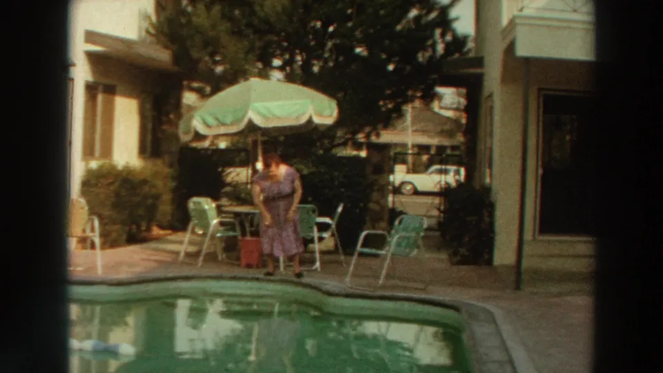 1966 Puerto Rico frame showing a woman bending at the pool edge near metal patio chairs, umbrella shade, and a motel exterior walkway.
