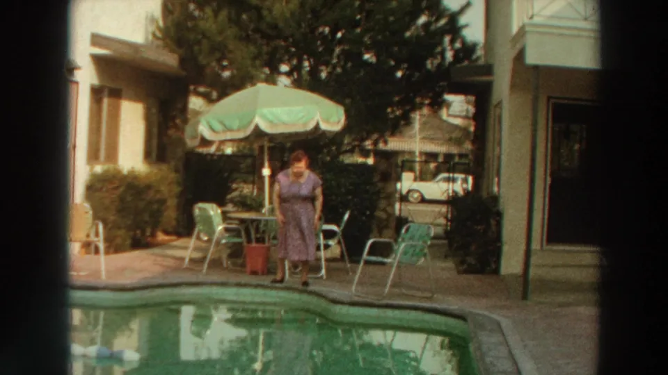1966 Puerto Rico home-movie frame of a woman by the motel pool, with patio furniture, a green-and-white umbrella, and a white car in the background.