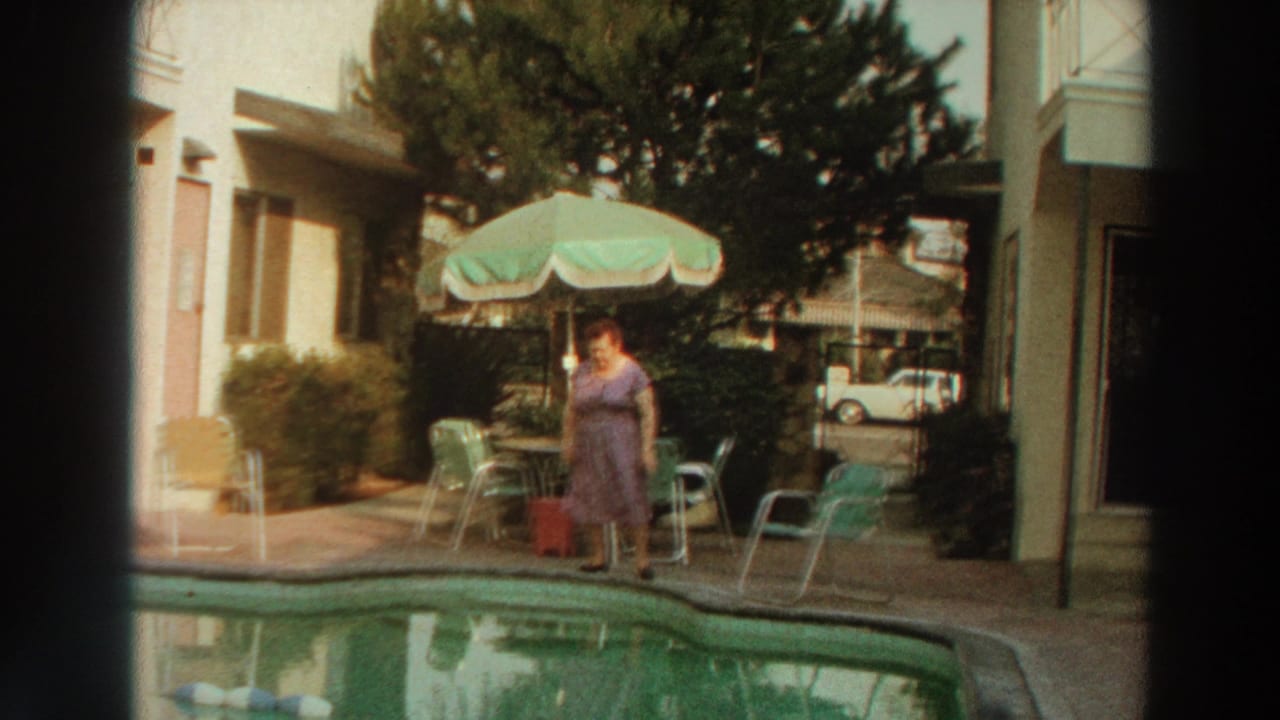 Woman in a purple dress stands near a swimming pool with a green umbrella and patio chairs behind her.