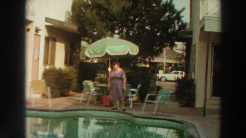 In 1966 Puerto Rico home-movie footage, a woman in a purple dress stands by a pool near a green umbrella, metal chairs, and a low building.