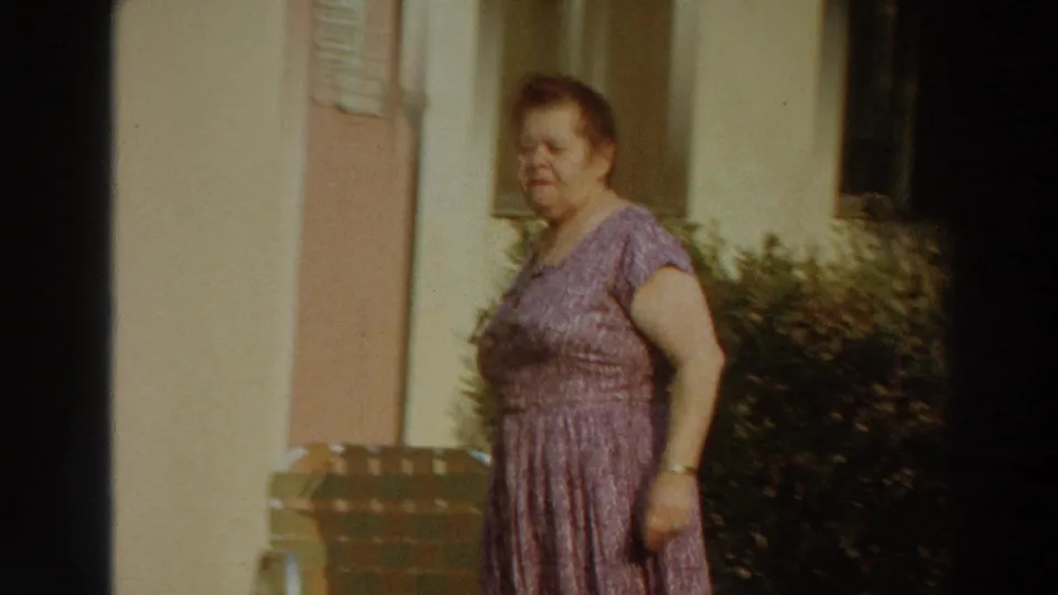 A woman in a purple patterned dress walks past a sunlit building exterior in Puerto Rico, with shrubs and poolside furniture nearby.