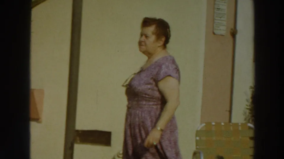 Side view of the woman walking past a light-colored exterior wall, with low outdoor furniture and a posted sign partly visible.