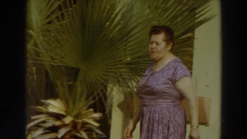 The woman continues past palm foliage and a light building wall, maintaining the quiet poolside movement seen in the 1966 Puerto Rico footage.