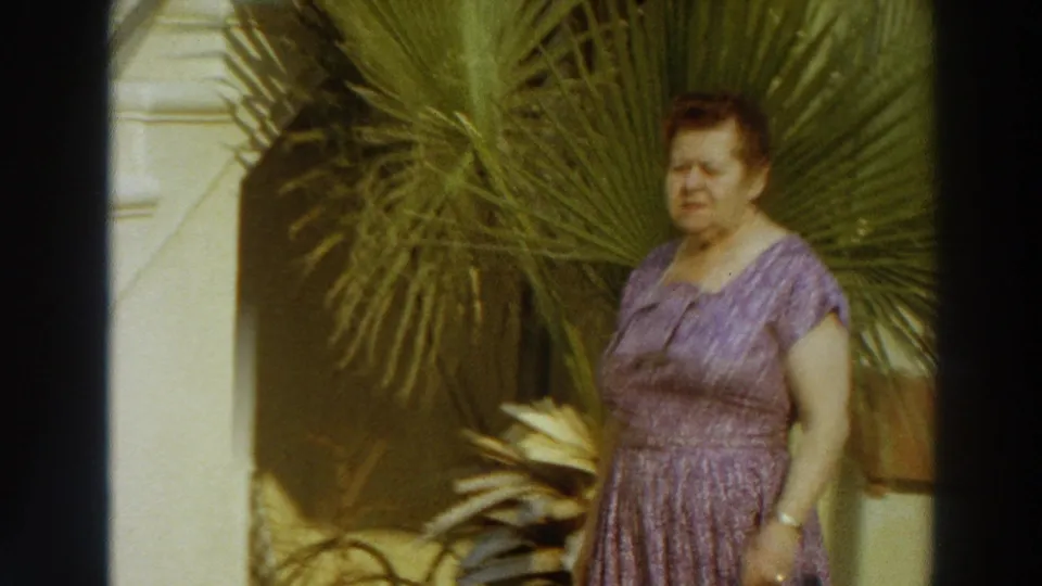 In Puerto Rico home-movie footage from 1966, a woman in a lilac patterned dress stands beside palm fronds in bright sun.