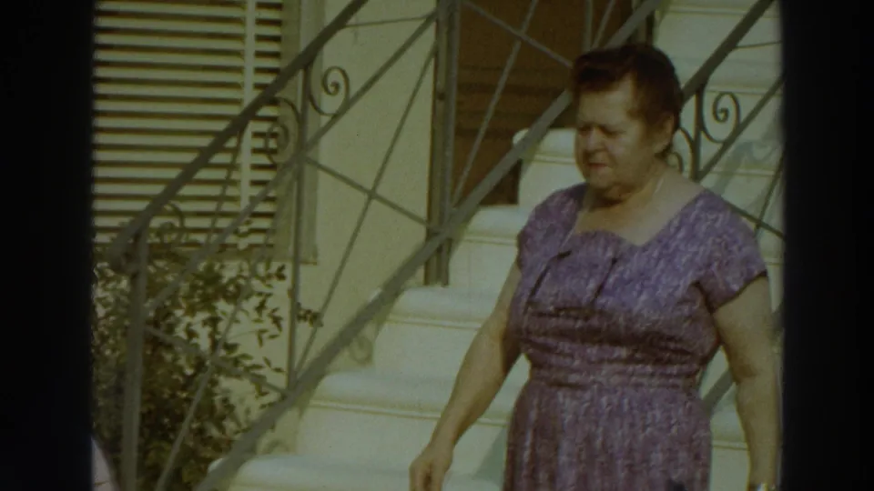 A woman in a lilac patterned dress moves past shutters, stairs, plants, and metal railings in Puerto Rico, 1966.