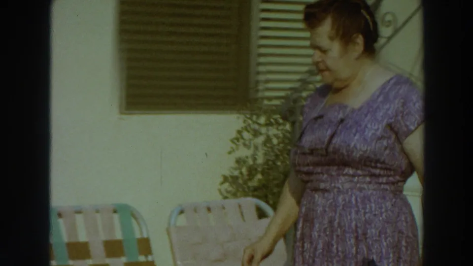 The woman continues along a sunlit exterior wall with patio chairs, shutters, and plants visible in Puerto Rico, 1966.