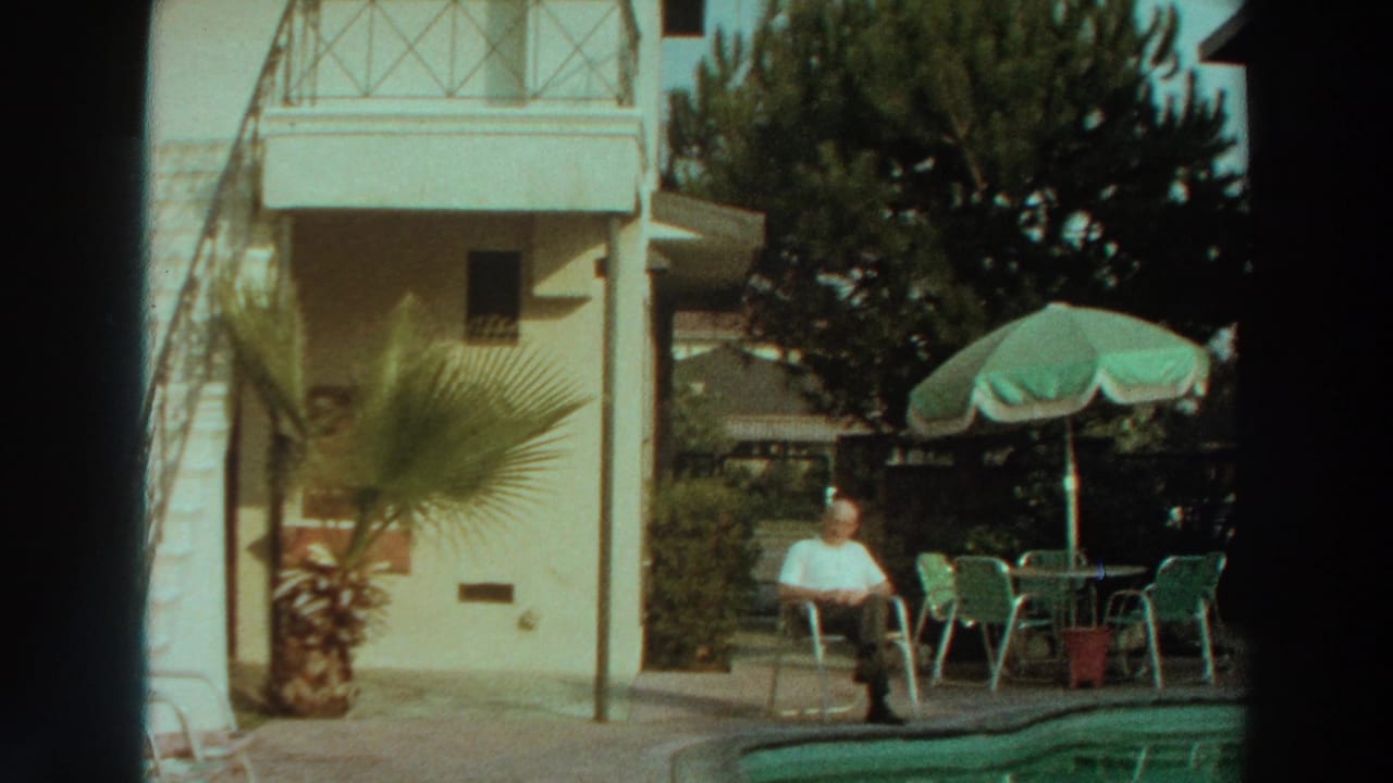 Man seated by a swimming pool near a green umbrella and patio furniture.