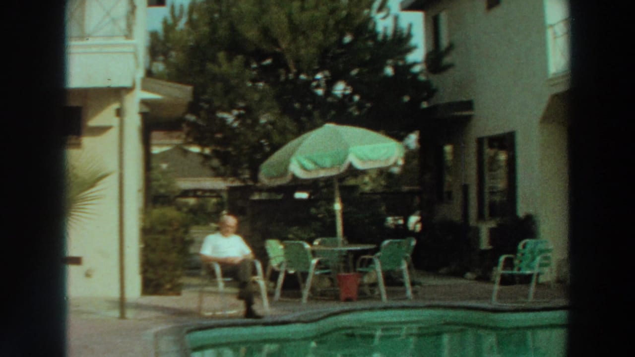 Man in a white shirt sits beside a pool with patio chairs and an umbrella behind him.