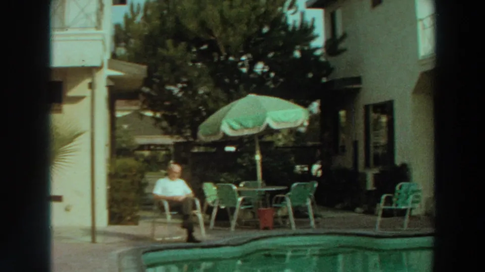 A 1966 Puerto Rico poolside view shows a man seated in the sun near green patio chairs, an umbrella, trees, and a pale building.