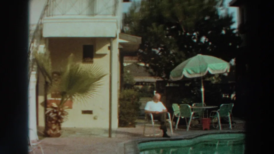 A man sits beside a curved swimming pool in Puerto Rico in 1966, with a green umbrella, patio chairs, plants, and a light building visible.