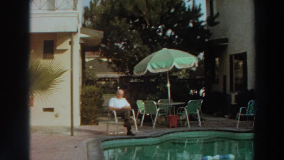 1966 Puerto Rico home-movie frame showing an elderly man seated near a pool with patio furniture and tropical plants.