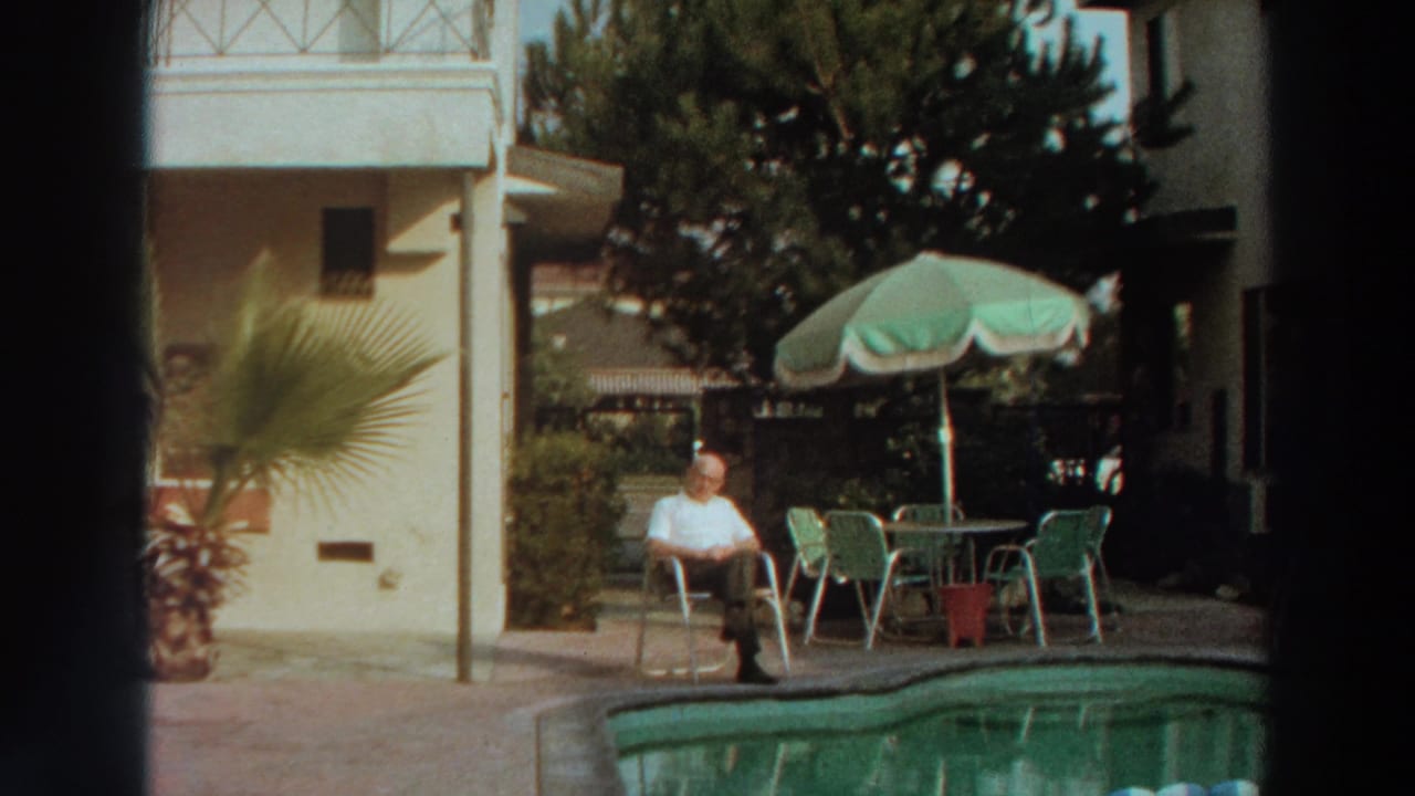 1960s 8mm frame of a man seated by a swimming pool with outdoor furniture, plants, and a light-colored building behind him.