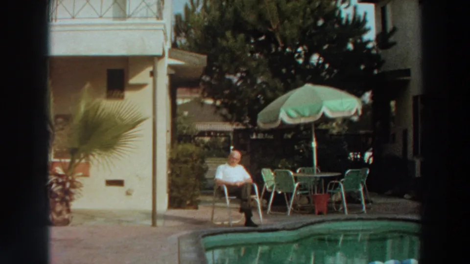 1960s 8mm frame of a man seated by a swimming pool with outdoor furniture, plants, and a light-colored building behind him.