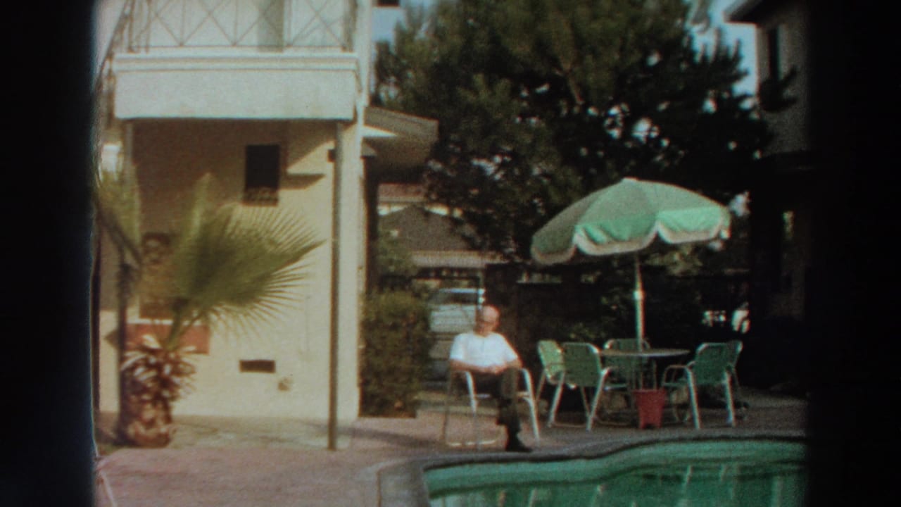 Puerto Rico poolside home-movie frame with an elderly man seated near a green umbrella, outdoor chairs, and sunlit foliage.
