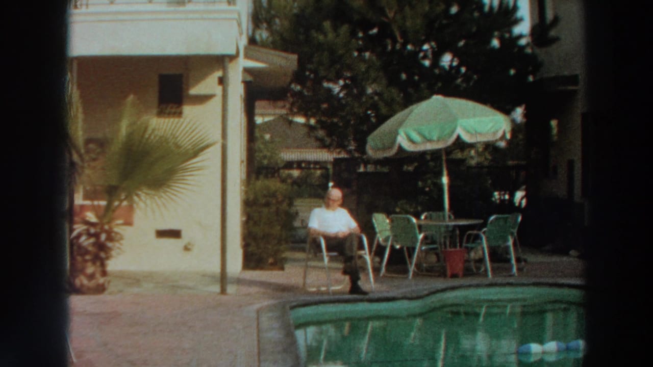 Man in a white shirt seated beside a backyard pool under a green patio umbrella.
