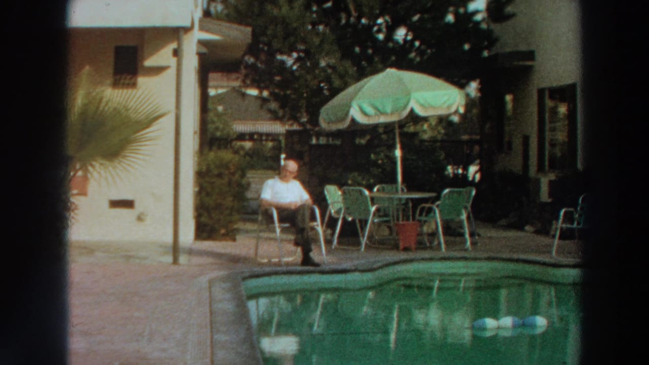 Man seated beside a swimming pool near a green parasol and patio chairs.