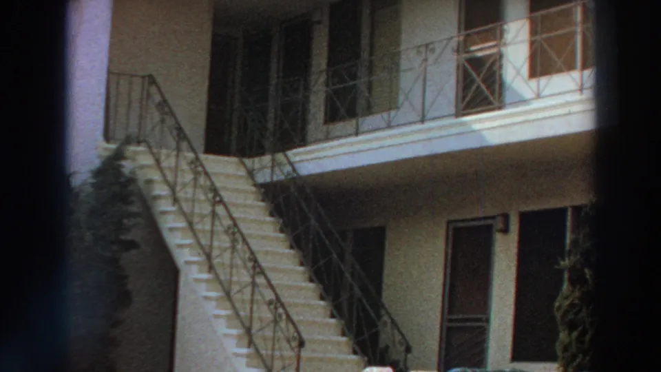 1966 Puerto Rico home-movie frame of a light residential exterior with stairs, railings, shaded doors and a partial plant at the edge.