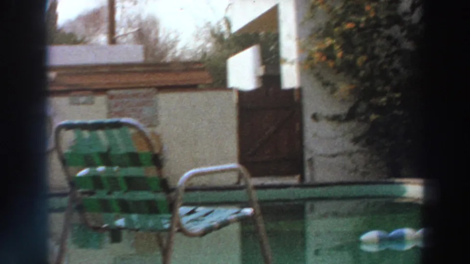 1966 Puerto Rico poolside frame with striped lawn chair, metal chair, green water, stucco wall, and wooden gate.