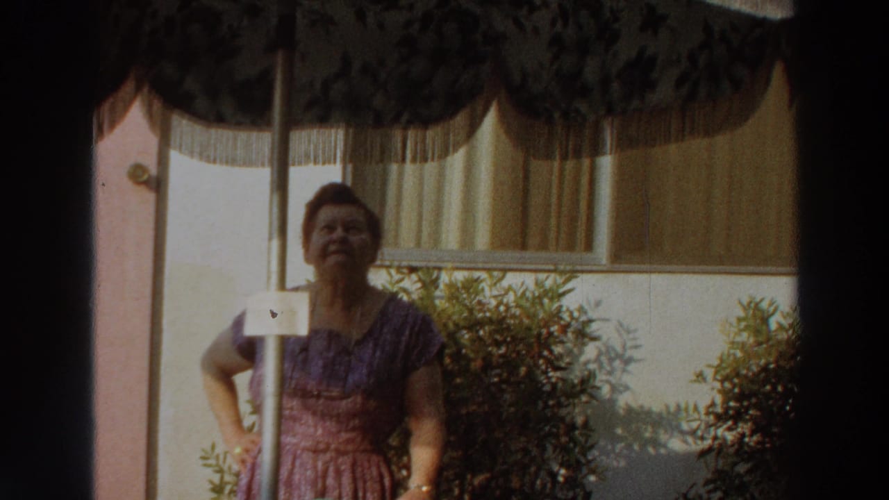 Woman in a purple dress stands under a patio umbrella beside shrubs and a curtained window.