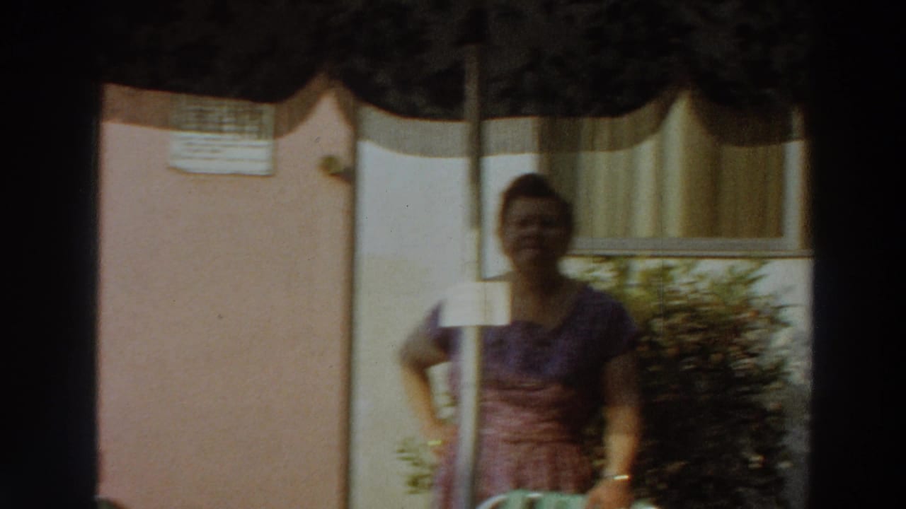The woman remains posed in the sunny backyard, partly shaded by the umbrella, with the exterior wall, window curtains, and shrubbery visible.