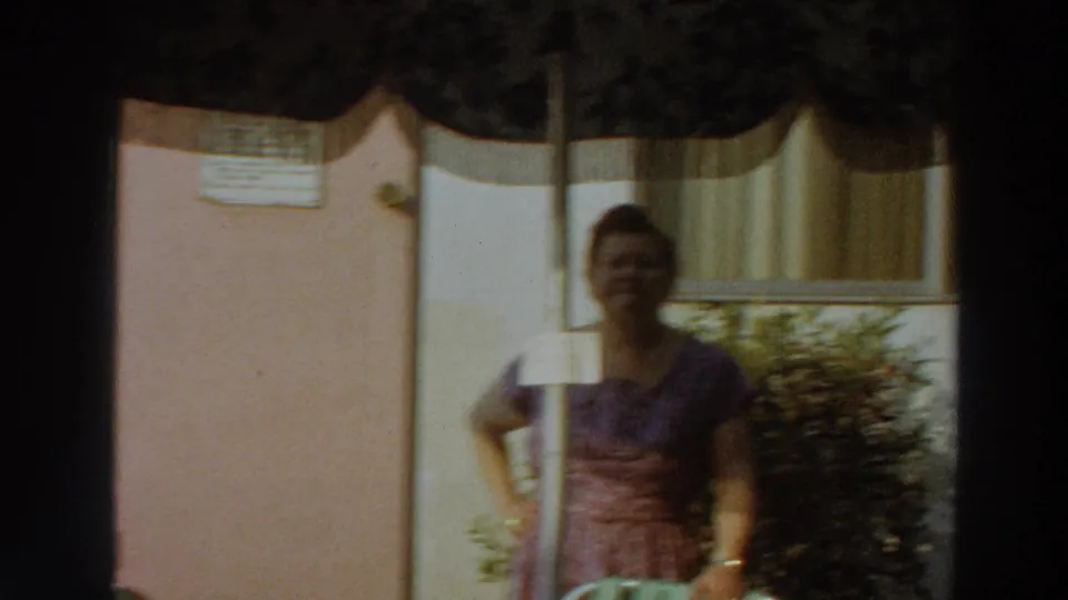 The woman remains posed in the sunny backyard, partly shaded by the umbrella, with the exterior wall, window curtains, and shrubbery visible.