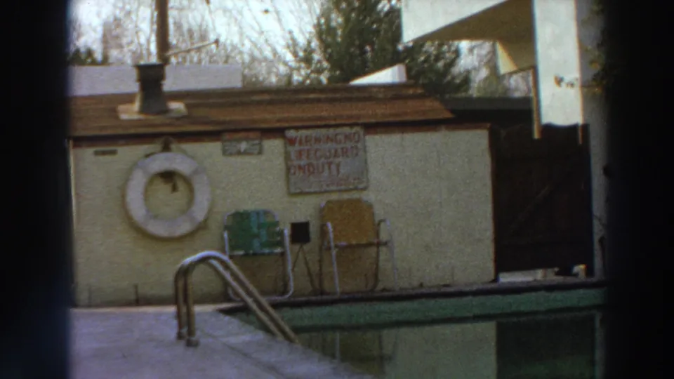 1966 Puerto Rico home-movie frame showing a poolside hut with a life ring, chairs, a warning sign, and the edge of a swimming pool.