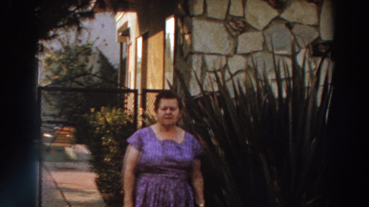 1966 Puerto Rico home-movie frame of an adult woman in a purple patterned dress standing outside a stone-faced building near tall plants.