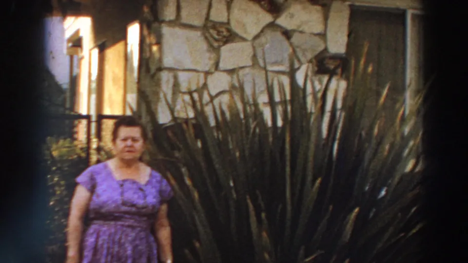 Adult woman in a purple patterned dress stands near dense tall plants beside a stone-faced exterior in 1966 Puerto Rico footage.