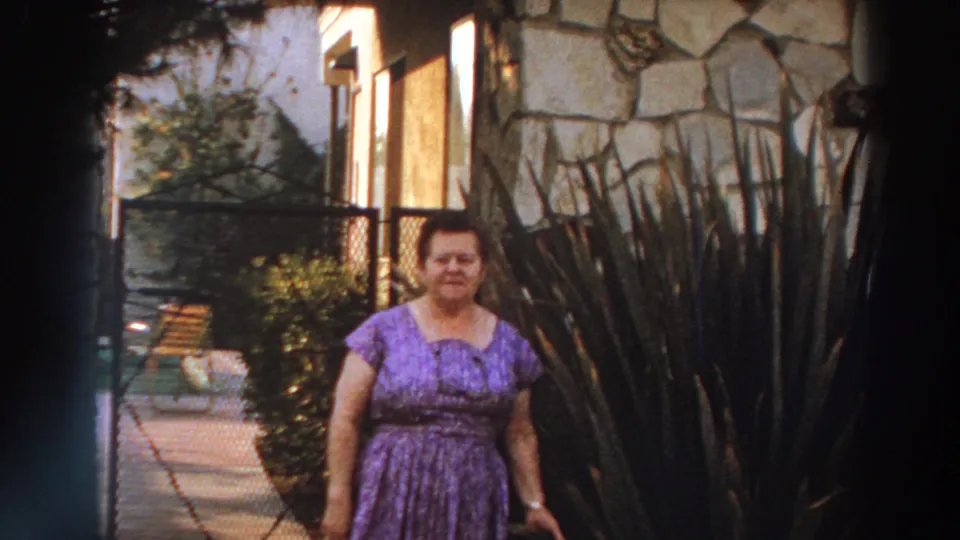 1966 Puerto Rico home-movie frame showing the woman facing the camera near a stone wall, walkway, and tall landscaping plants.