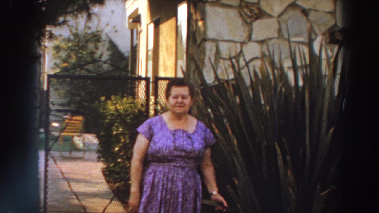 In Puerto Rico home-movie footage from 1966, a woman in a purple patterned dress stands near a stone house wall, gate and tall garden plants.