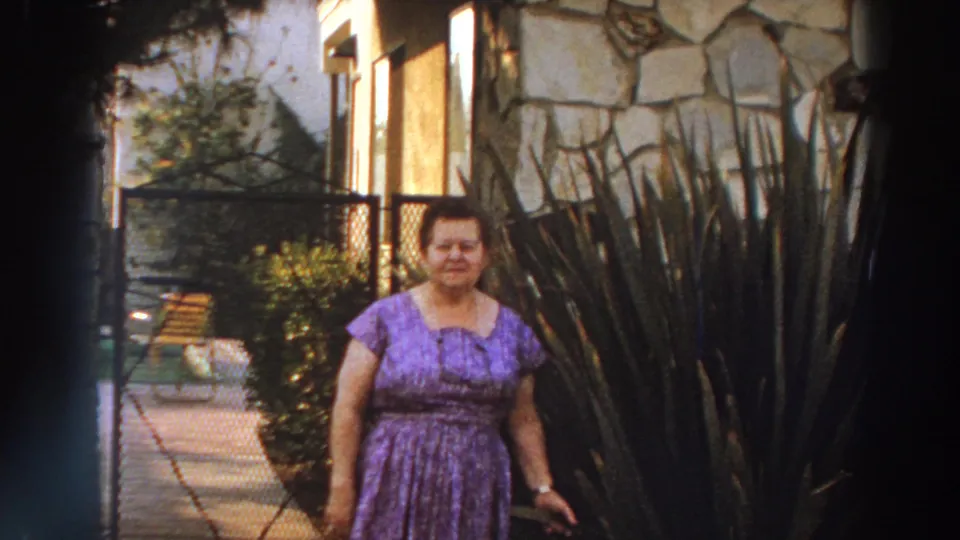 In Puerto Rico home-movie footage from 1966, a woman in a purple patterned dress stands near a stone house wall, gate and tall garden plants.