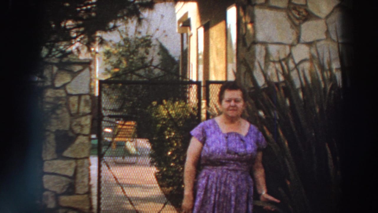 The woman remains posed in the garden beside tall plants, with a stone wall, chain-link gate and patio furniture visible in the 1966 Puerto Rico scene.