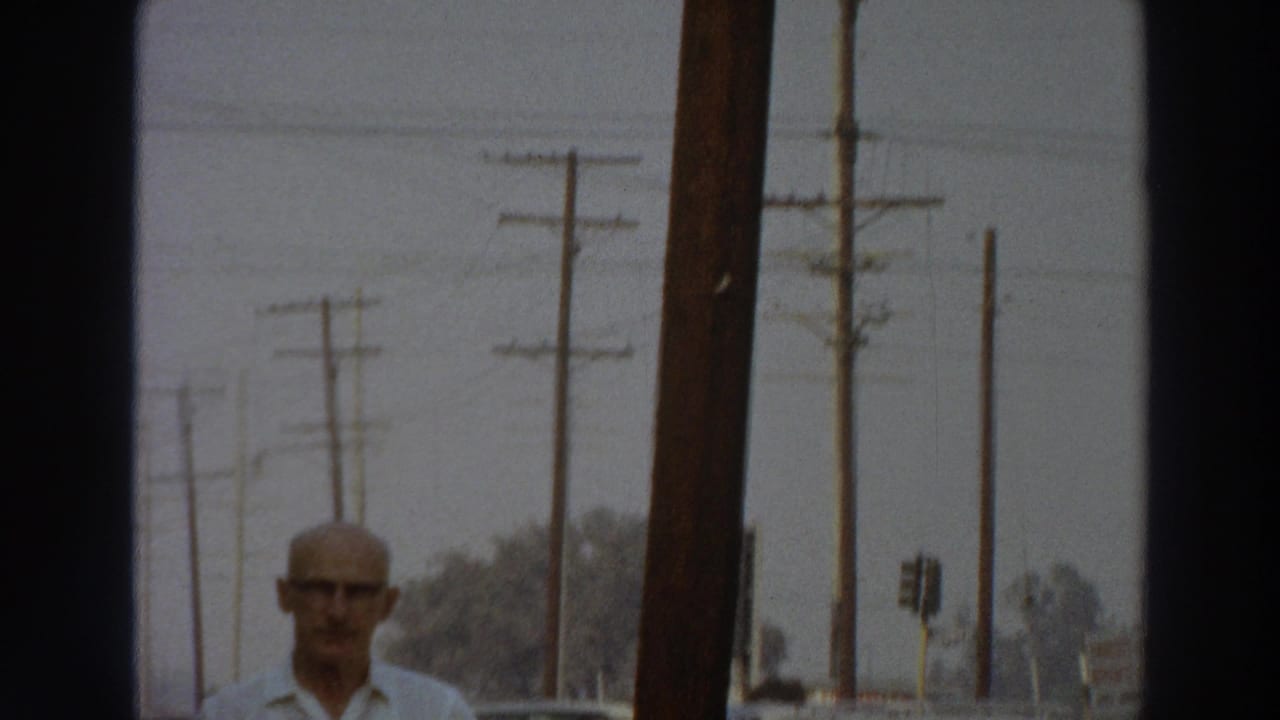 1966 Puerto Rico 8mm frame showing a bald man with glasses entering view beside a sidewalk, parked cars, and utility poles.