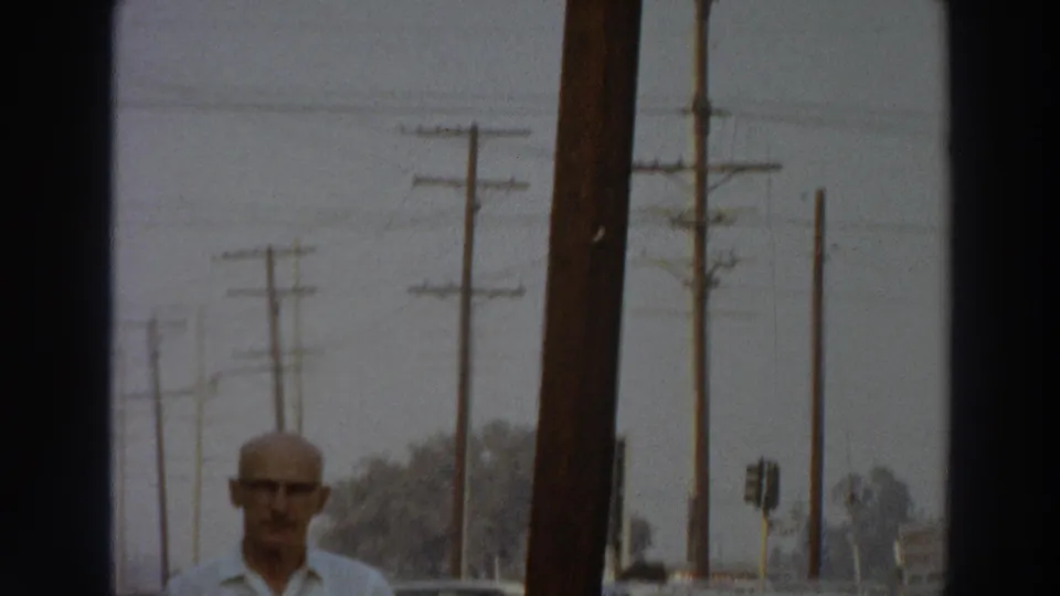 1966 Puerto Rico 8mm frame showing a bald man with glasses entering view beside a sidewalk, parked cars, and utility poles.