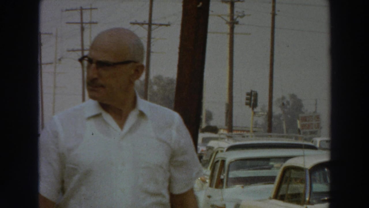 The man glances to the side while passing the camera on a 1966 Puerto Rico sidewalk lined with parked cars.
